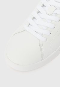 Krupan plan bijele kožne tenisice s bijelim vezicama i vidljivim šavovima na svijetloj pozadini.