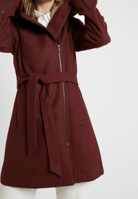 Cappotto in misto lana bordeaux con cappuccio, presenta una chiusura frontale con zip, vita regolabile con cintura e due tasche frontali. Taglio dritto classico e texture morbida.