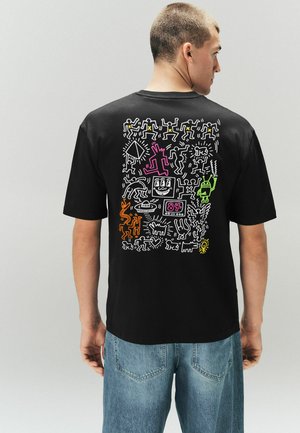 Mężczyzna w czarnej koszulce z kolorowymi, abstrakcyjnymi rysunkowymi postaciami i kształtami na plecach, w połączeniu z niebieskimi dżinsami.