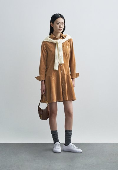 Καφέ φόρεμα πουκαμίσας από κορδούρα με φαρδιά βάση, μπροστινό κουμπί και μακριά μανίκια. Λευκό πλεκτό πουλόβερ δεμένο γύρω από το λαιμό. Γκρι κάλτσες και λευκά αθλητικά παπούτσια.