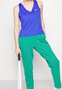 Top deportivo morado con escote en V y logo blanco, combinado con pantalones de chándal verdes. El modelo sostiene una raqueta de tenis blanca.