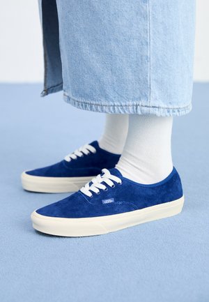 Baskets Vans en daim bleu avec des lacets blancs, portées avec des chaussettes blanches et un pantalon en denim bleu clair, sur une surface bleue.