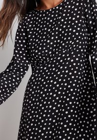Schwarzes Kleid mit weißen Punkten, langen Ärmeln, rundem Ausschnitt und Raffung in der Taille. Glatter, leichter Stoff.