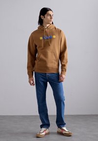 Hoodie en coton marron avec une poche avant et du texte graphique. Assorti à un jean en denim bleu et des baskets multicolores. Fond simple.