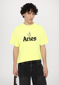 T-shirt oversize giallo neon con una grafica di un'anatra cartone e il testo "Aries" in grassetto nero. Indossato con jeans scuri.
