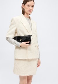 Γυναίκα που φοράει υφασμάτινο εκρού σακάκι και φούστα, κρατώντας μαύρο τσαντάκι πατέντα, σε ανοιχτό γκρι φόντο.