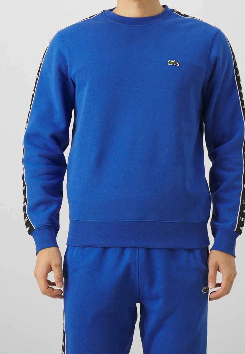 Sweat-shirt bleu en tissu doux, doté d'un col rond côtelé et de bandes latérales noires avec texte blanc. Logo Lacoste sur la poitrine.