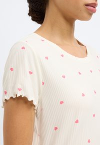 Κοντομάνικο κρεμ τοπ με ριμπ υφή και μικρά ροζ μοτίβα καρδιάς. Διαθέτει στρογγυλεμένο λαιμόκοψη και βολάν στις άκρες των μανικιών.