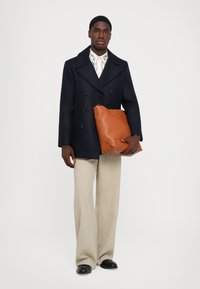 Muž v námorníckom dvojradovom kabáte, vzorovanej bielej košeli, béžových širokých nohaviciach, čiernych topánkach, s veľkou hnedou koženou taškou.