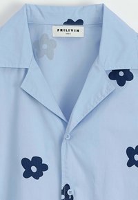 Chemise bleu clair à manches courtes, ornée de motifs floraux bleu foncé, avec un col pointu et deux boutons à l'avant. Étiquette indiquant "FRILIVIN, taille M."