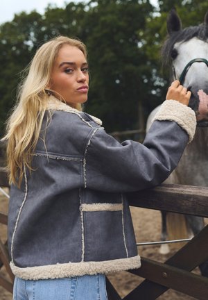 Grå faux läderjacka med vit fleecefoder och manschetter, med en boxig design och framficka, bärs av en person nära en häst.