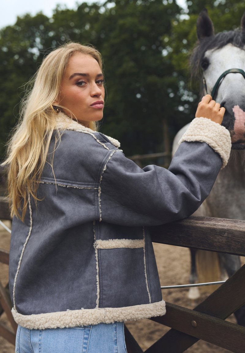 Szara kurtka z sztucznej skóry z białym futerkiem wewnątrz i przy mankietach, o pudełkowym kroju, z kieszenią z przodu, noszona przez osobę w pobliżu konia.