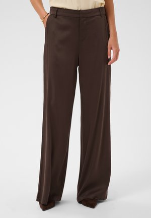 Personne portant un pantalon marron foncé taille haute, à jambes larges avec passants de ceinture, associé à des chaussures pointues foncées sur un fond uni.