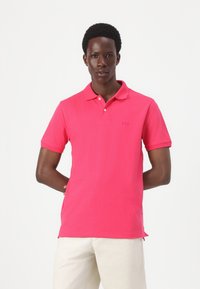 Fuchsia poloshirt med krave, korte ærmer og lille logo på brystet. Modellen har lysfarvede bukser på. Glat stofstruktur.
