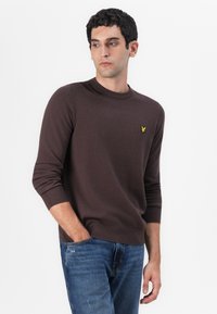Pull en tricot marron avec un col rond et des poignets côtelés. Présente un petit logo jaune sur la poitrine. Associé à un jean bleu.