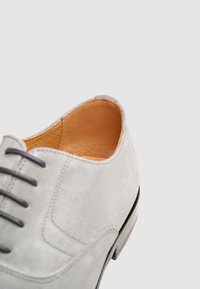 Chaussure en daim gris avec un bout arrondi, dotée de lacets noirs et d'un intérieur en cuir. La semelle extérieure est légèrement surélevée avec un accent noir.