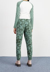 Person som bär gröna och mörkgrå mönstrade joggers med elastiska muddar, en vit och grön långärmad tröja och beige stövlar, som står på en matta.