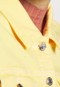 Giacca di jeans gialla con colletto a punta, bottoni a pressione argentati e cuciture bianche, indossata sopra un top rosa a coste.
