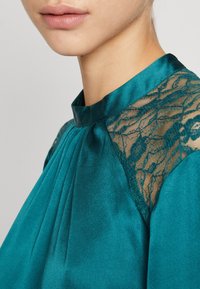 Haut en satin bleu sarcelle avec plis, doté de détails en dentelle noire transparente sur les épaules et d'un col mandarin. Texture lisse et design élégant.