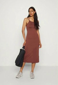 Vestido midi castanho sem mangas feito de um tecido suave, com um design ajustado. A modelo traz uma bolsa preta e usa tênis claros.