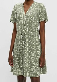 Robe à manches courtes en vert olive, avec un décolleté en V, un motif à pois blancs, un devant boutonné et une taille nouée pour un ajustement réglable.