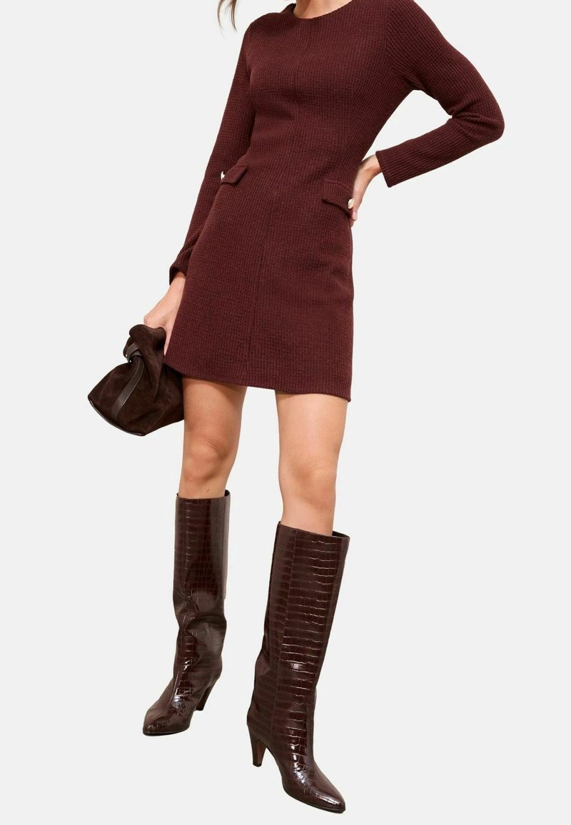 Femme portant une robe ajustée à manches longues de couleur bordeaux avec deux poches avant, tenant une pochette assortie bordeaux et portant des bottes à texture montant jusqu'aux genoux.