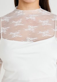 Robe blanche avec un haut en dentelle transparente ornée de motifs floraux. Matière lisse en dessous, avec un col rond et une silhouette ajustée.