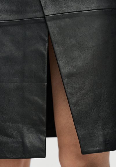 Μαύρη δερμάτινη φούστα με μπροστινή σχισμή που αποκαλύπτει γυμνά πόδια φορεμένη από ένα άτομο, εμφανίζοντας λεπτομέρειες υφής και ραφής στη φούστα.