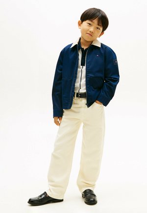 Jongen staat met handen in zakken, draagt een navy jas, gestreept shirt, beige broek en zwarte loafers, tegen een effen witte achtergrond.