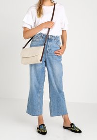 Kvinna klädd i vit t-shirt, ljusblå vida jeans, svarta broderade slip-on skor och en beige axelväska med mörk rem.