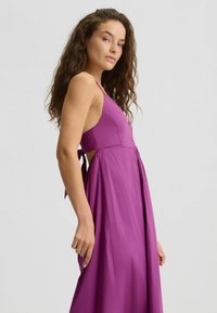 Purple sleeveless dress with a fitted bodice and flowing skirt. Features side cutouts and a tied waist detail, made from smooth fabric.