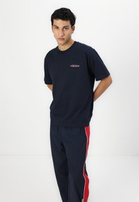 Tamsiai mėlynas crew-neck marškinėliai su trumpomis rankovėmis, ant kurių yra mažas "Guess Jeans" logotipas. Derinamas su tamsiai mėlynomis kelnėmis, papuoštomis raudonomis šoninėmis juostelėmis.