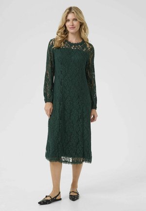Green lace dress with long sleeves, knee-length hem, floral pattern, and scalloped details at the neckline and hem; worn with black flats.