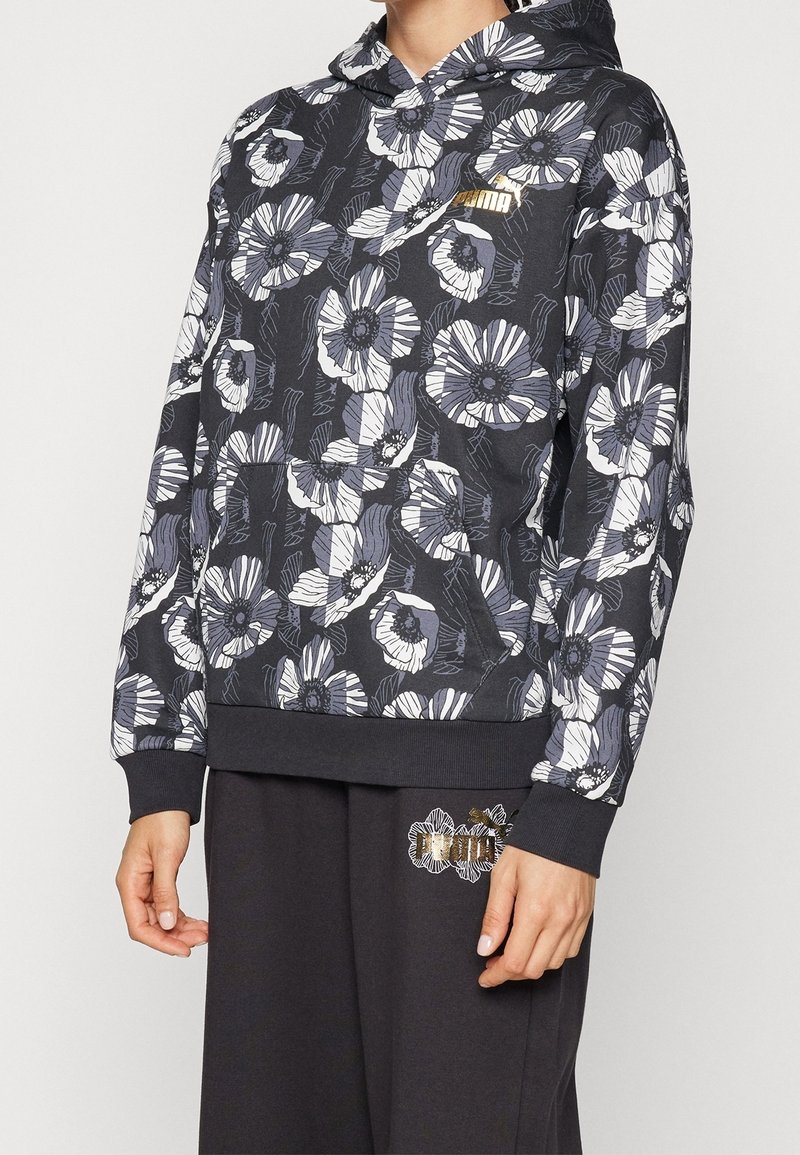 Person in einem schwarzen Kapuzenpullover mit floralem Muster und goldener Puma-Logo sowie passenden schwarzen Hosen mit floralem Puma-Design.