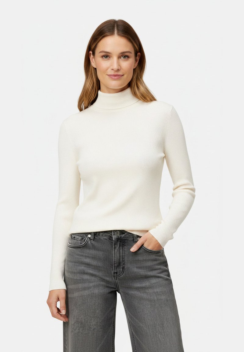 Femme aux cheveux châtain clair portant un pull à col roulé blanc et un jean gris, une main dans une poche, sur un fond uni.