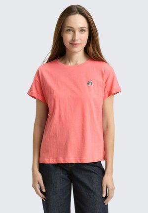 Junge Frau mit langen braunen Haaren, die ein korallenfarbiges T-Shirt mit kurzen Ärmeln und einem kleinen schwarz-weißen Blattlogo trägt, kombiniert mit dunkelblauen Jeans.