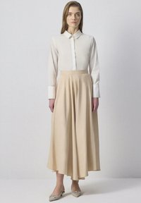 Woman standing against plain background wearing a white buttoned shirt, beige asymmetrical midi skirt, and beige pointed flats with embellishments.