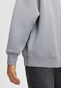 Braccio sinistro di una persona che indossa una felpa grigia chiara con etichetta nera "Strellson", abbinata a jeans grigi scuri, su sfondo neutro.