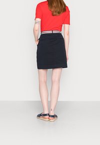 Gonna di jeans blu navy con passanti per cintura e cintura in tessuto a righe, abbinata a una camicia rossa aderente e sandali espadrillas. Presenta tasche e un taglio a lunghezza ginocchio.