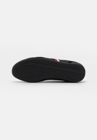 Semelle de basket noire avec un motif géométrique texturé et dessus noir comportant une bande rouge, blanche et bleue sur fond blanc.