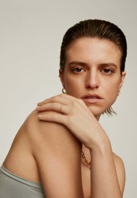 Anillo de oro con pequeñas piedras en una mano que descansa sobre un hombro. Textura de piel suave, camiseta gris neutro y un collar de cuentas simple.