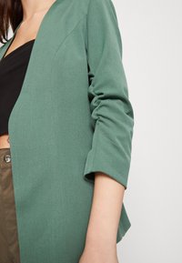 Grön blazer med en mjuk textur, öppen fram och trekvartslånga ärmar. Har subtil sömnad och en avslappnad passform, tillsammans med en svart topp.