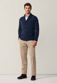 Cardigan di lana navy con colletto a scialle e chiusura con bottoni, abbinato a pantaloni beige e scarpe in camoscio marrone scuro. Texture liscia, design aderente.