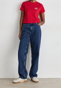 Röd t-shirt med kort ärm och vitt logo, kombinerad med blå högmidjade raka jeans och ljusa sneakers.