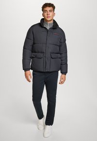 Giacca a vento nera con colletto alto, chiusura a bottoni, due tasche frontali con pattina e una consistenza morbida. Abbinata a pantaloni scuri e sneakers bianche.