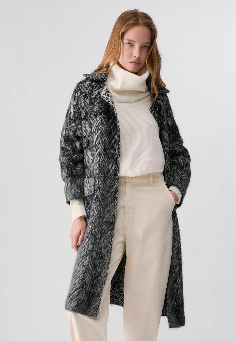 Jeune femme portant un long manteau noir et gris à motifs par-dessus un pull à col roulé blanc et un pantalon beige, debout devant un fond uni.