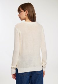 Donna con capelli castani lisci che indossa un maglione di lana avorio e jeans blu, di spalle su uno sfondo chiaro e uniforme.