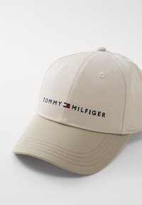 Gorra de béisbol beige hecha de algodón. Presenta el logotipo bordado de "TOMMY HILFIGER" en negro con acentos en rojo y azul. Visera curva, diseño de seis paneles.