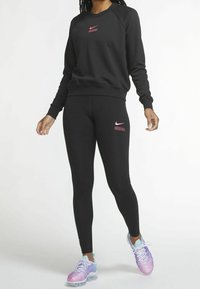 Svart Nike-tröja med rosa logotyp, kombinerad med svarta leggings med rosa accenter. Modellen bär iriserande sneakers. Slät tygtextur.