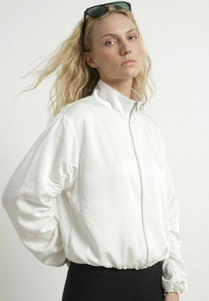 Blond kvinde med solbriller på hovedet bærer hvid lynlåstrøje og sorte bukser, stående mod en ensfarvet lys baggrund.